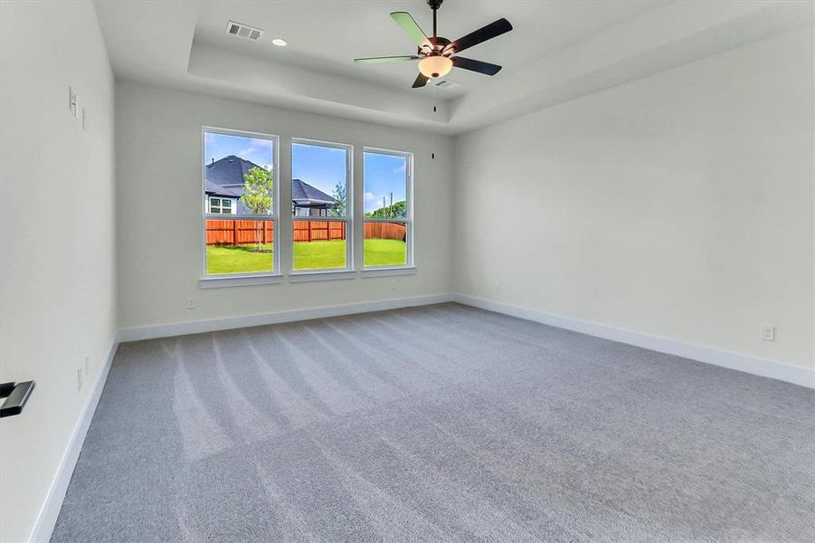 Spacious room featuring light gray carpeting, a tray ceiling with a ceiling fan, and three large windows overlooking a fenced backyard with green lawn Spacious room featuring light gray carpeting, a tray ceiling with a ceiling fan, and three large windows overlooking a fenced backyard with green lawn