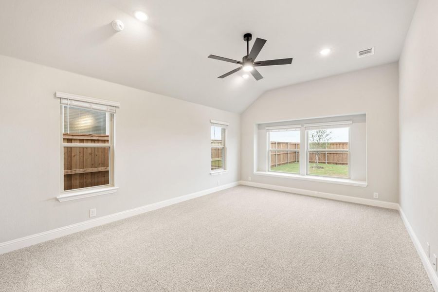Empty bedroom with vaulted ceiling, ceiling fan, carpet flooring, and multiple windows overlooking backyard