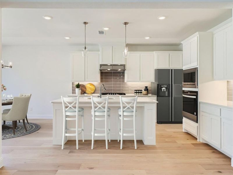 Virtually staged photo - Kitchen featuring light wood-type flooring, wall chimney exhaust hood, appliances with stainless steel finishes, decorative backsplash, and pendant lighting