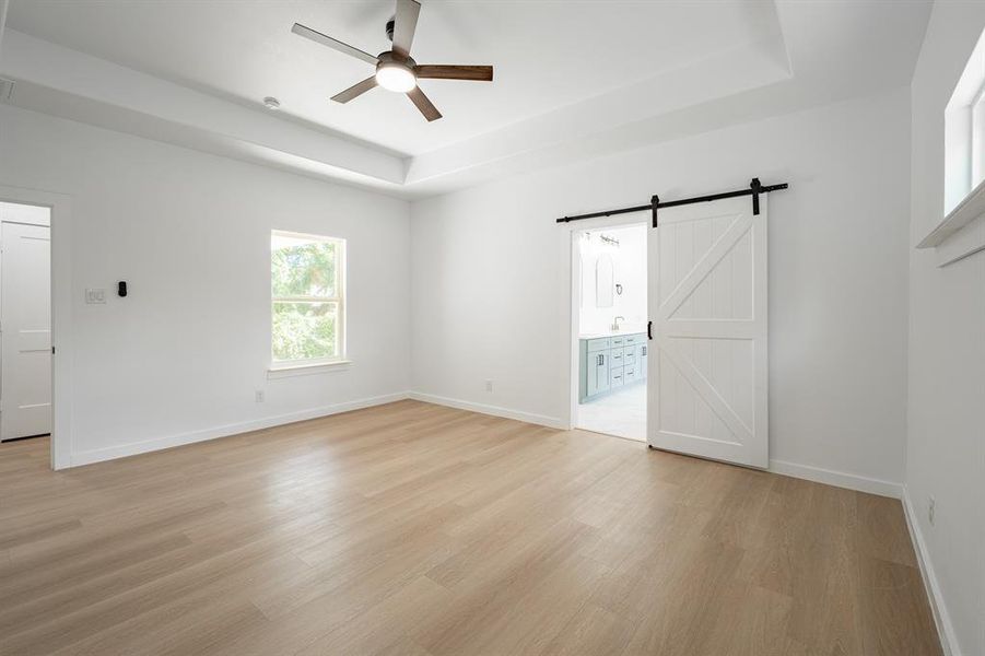 Unfurnished bedroom with a barn door, a raised ceiling, light wood finished floors, ensuite bath, and ceiling fan Unfurnished bedroom with a barn door, a raised ceiling, light wood finished floors, ensuite bath, and ceiling fan
