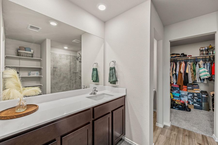 A well-lit primary bathroom vanity with a full-width mirror reflects a spacious shower and shelving.
