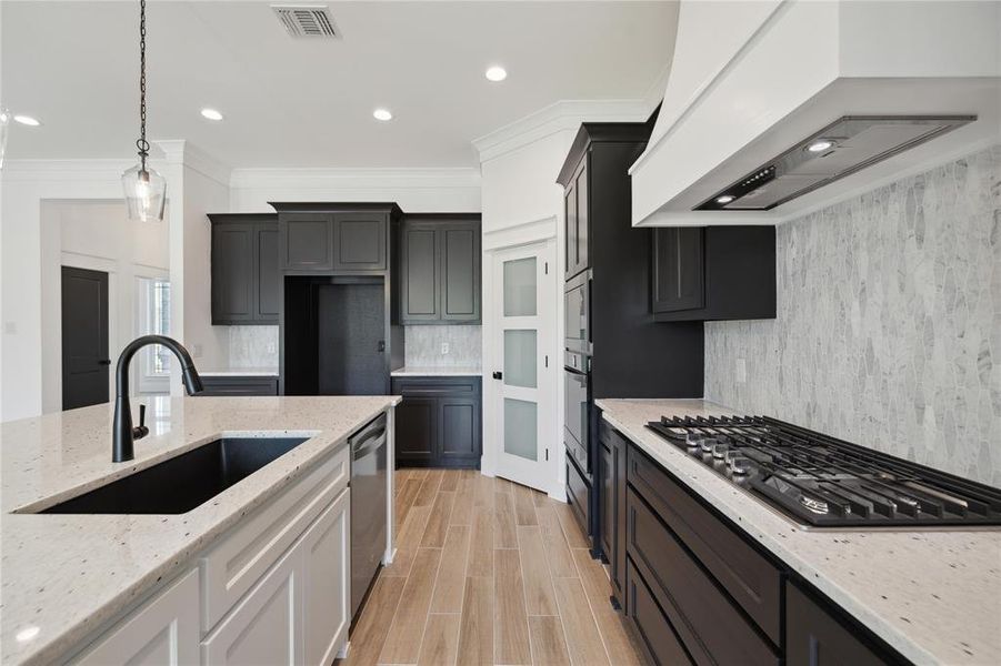 Kitchen with custom range hood, appliances with stainless steel finishes, crown molding, sink, and light stone counters