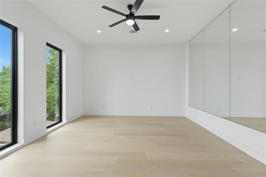 Spacious room with light wood-finish flooring, two large vertical windows, a ceiling fan with integrated lighting, recessed lighting, and a mirrored accent wall Spacious room with light wood-finish flooring, two large vertical windows, a ceiling fan with integrated lighting, recessed lighting, and a mirrored accent wall