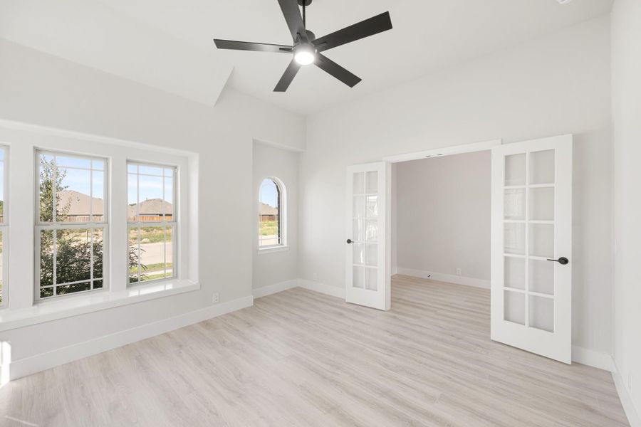 Empty room with light wood floors, ceiling fan, large windows, and white French doors