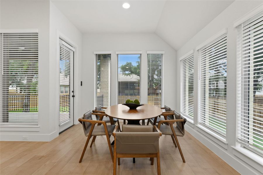 Vaulted ceiling dining area with wood-finish flooring, abundant natural light from multiple windows with blinds, and a glass-paned exterior door