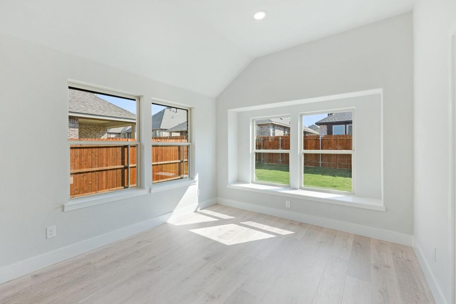 Empty bedroom with vaulted ceiling, light wood floors, and four windows overlooking backyard fence