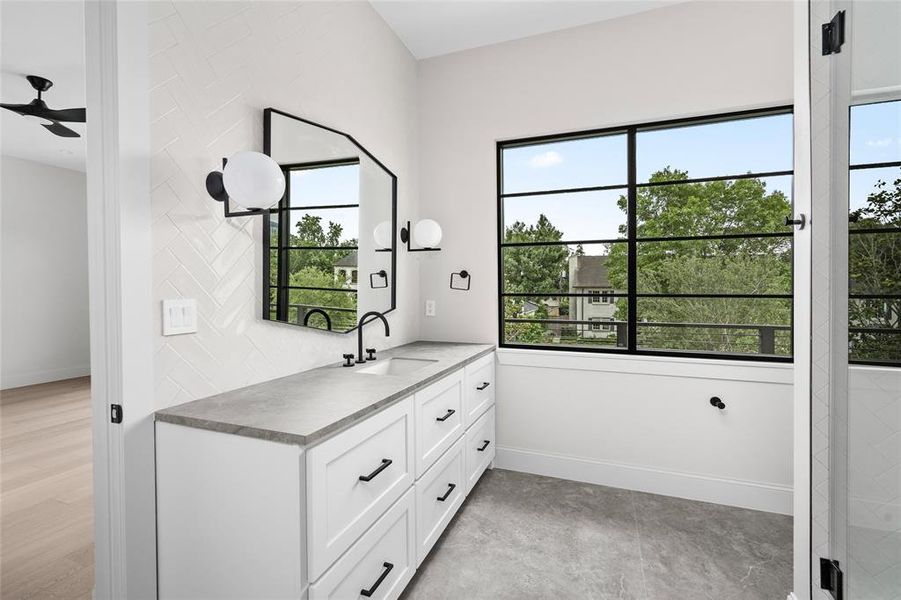 This ensuite bath boasts chic light fixtures, stone counters and flooring, a glass-enclosed tile walk-in shower, and an abundance of natural light.