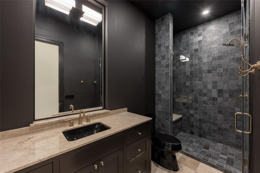 This striking full bathroom features a bold, moody palette with charcoal walls, a matching black toilet and sink, and natural stone countertops. The walk-in shower is wrapped in textured slate tile, paired with brass fixtures for a rich, dramatic finish. This striking full bathroom features a bold, moody palette with charcoal walls, a matching black toilet and sink, and natural stone countertops. The walk-in shower is wrapped in textured slate tile, paired with brass fixtures for a rich, dramatic finish.