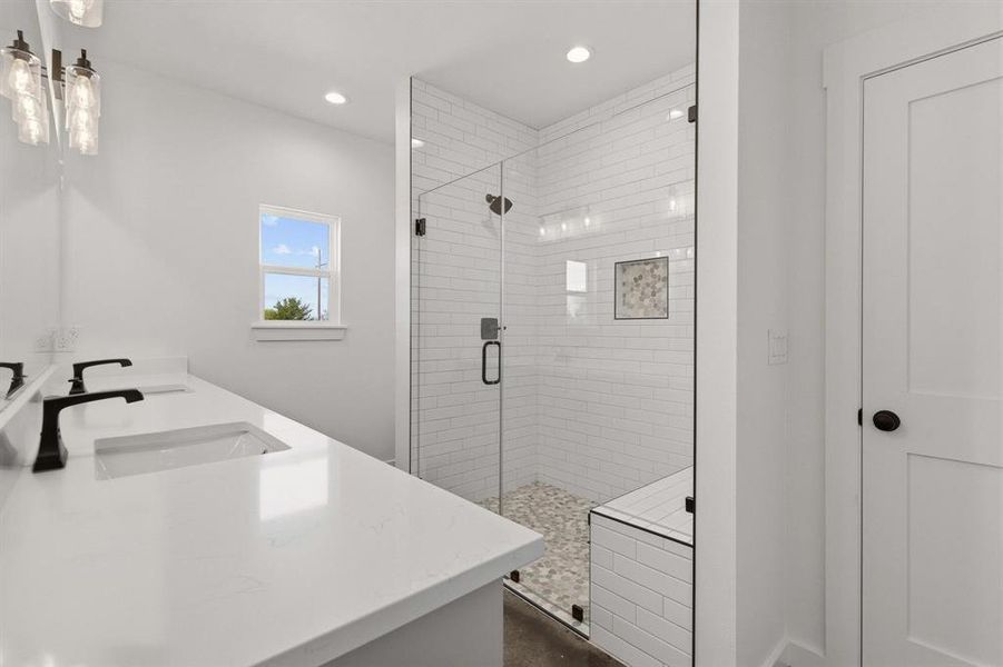Bathroom featuring a double vanity with matte black faucets, a tiled shower with a glass enclosure, a built-in shower bench, and a window providing natural light
