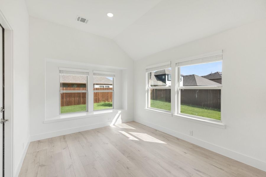 Empty bedroom with vaulted ceiling, light hardwood floors, and four windows overlooking backyard
