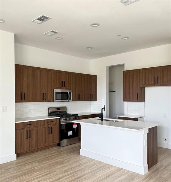 Kitchen with stainless steel appliances, tasteful backsplash, light countertops, light wood-style flooring, and an island with sink