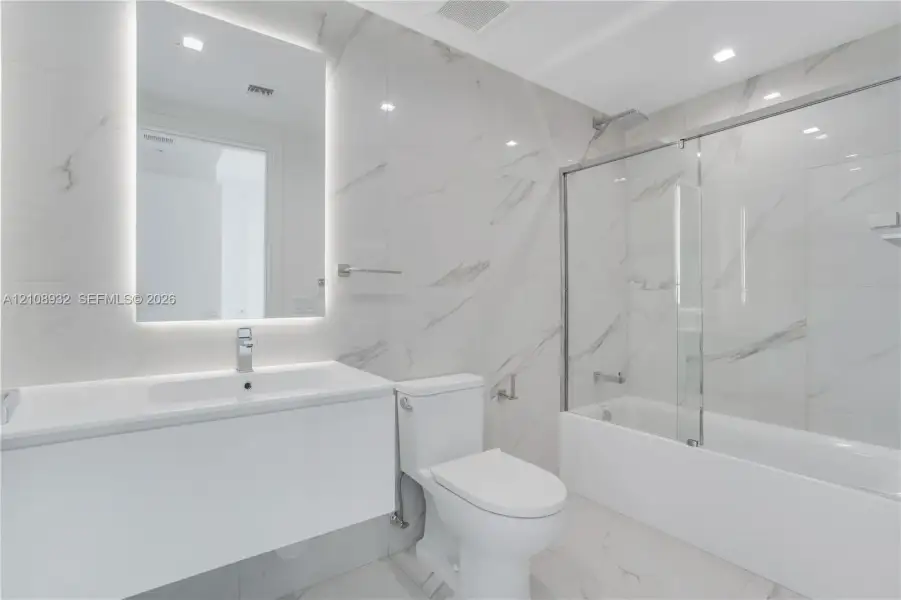 Bathroom featuring large-format stone-patterned wall and floor tile, a modern floating vanity with an integrated sink, a frameless glass tub enclosure, and an LED-backlit mirror