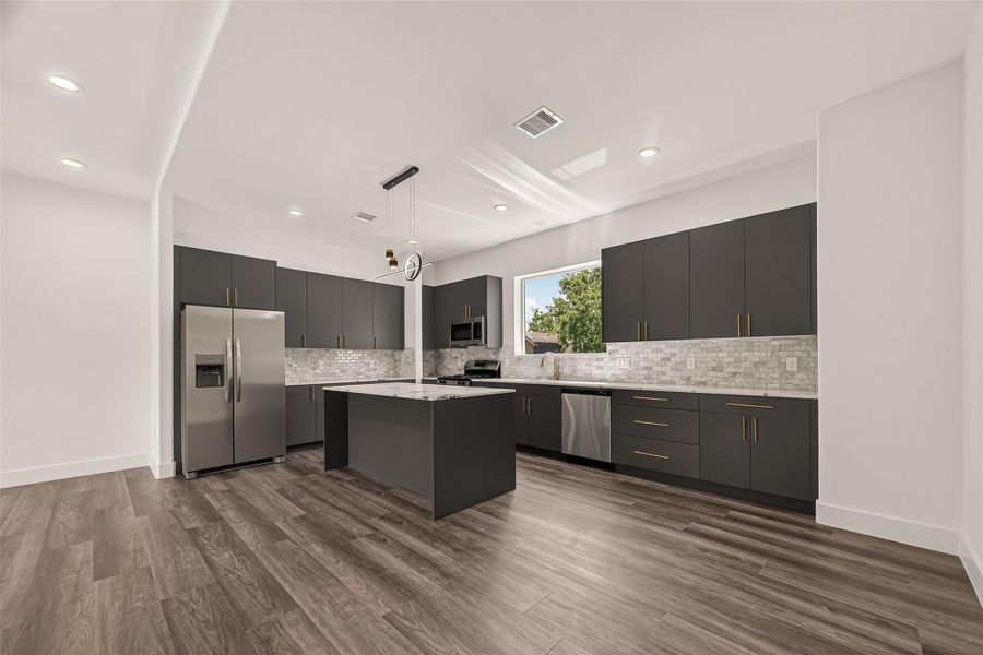 Modern kitchen with sleek dark cabinetry and stainless steel appliances. Features a central island with pendant lighting, tiled backsplash, and ample counter space. Bright and airy with vinyl flooring and large window. Modern kitchen with sleek dark cabinetry and stainless steel appliances. Features a central island with pendant lighting, tiled backsplash, and ample counter space. Bright and airy with vinyl flooring and large window.
