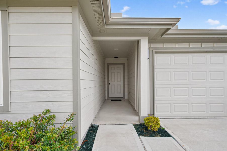 Modern entryway with clean lines, leading to a white front door. The exterior features light-colored siding and a neat pathway, with minimal landscaping for easy maintenance. Adjacent is a garage with a matching design.