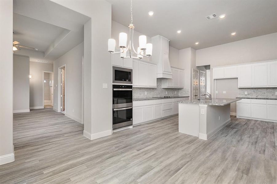 Kitchen with light stone counters, built in microwave, a kitchen island with sink, white cabinetry, and hanging light fixtures