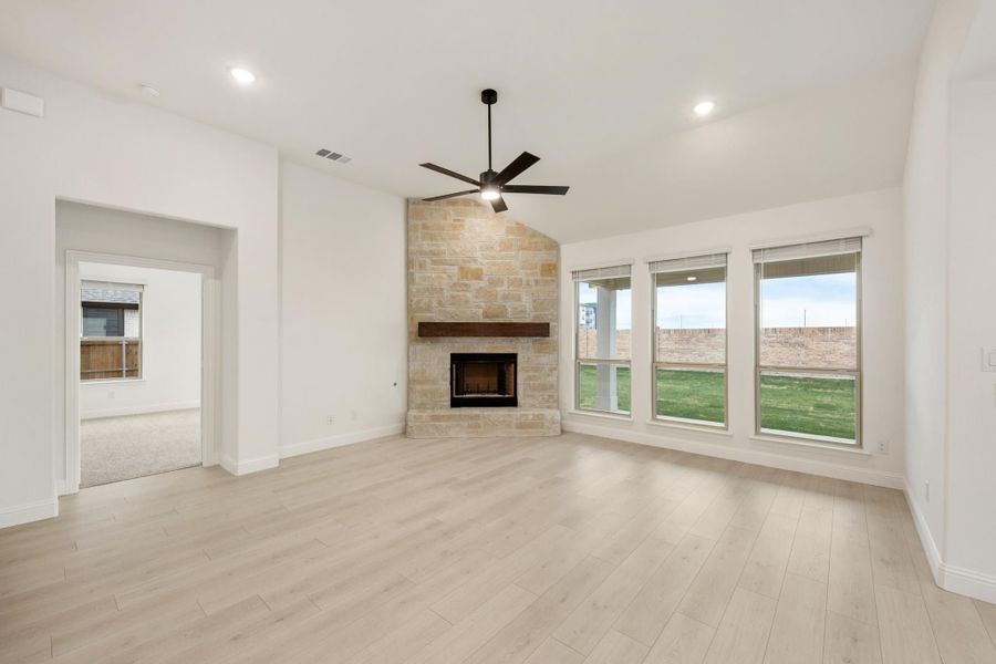 Open living room with stone fireplace, ceiling fan, light wood floors, and large windows Open living room with stone fireplace, ceiling fan, light wood floors, and large windows