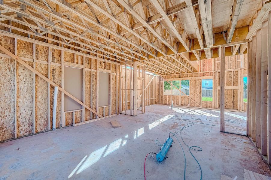 This photo shows a partially constructed room with exposed wooden framing and trusses, offering a glimpse of the structural layout and potential for customization. Large windows provide ample natural light, and there’s a view of the outdoors. Perfect for buyers looking to personalize their new home.
