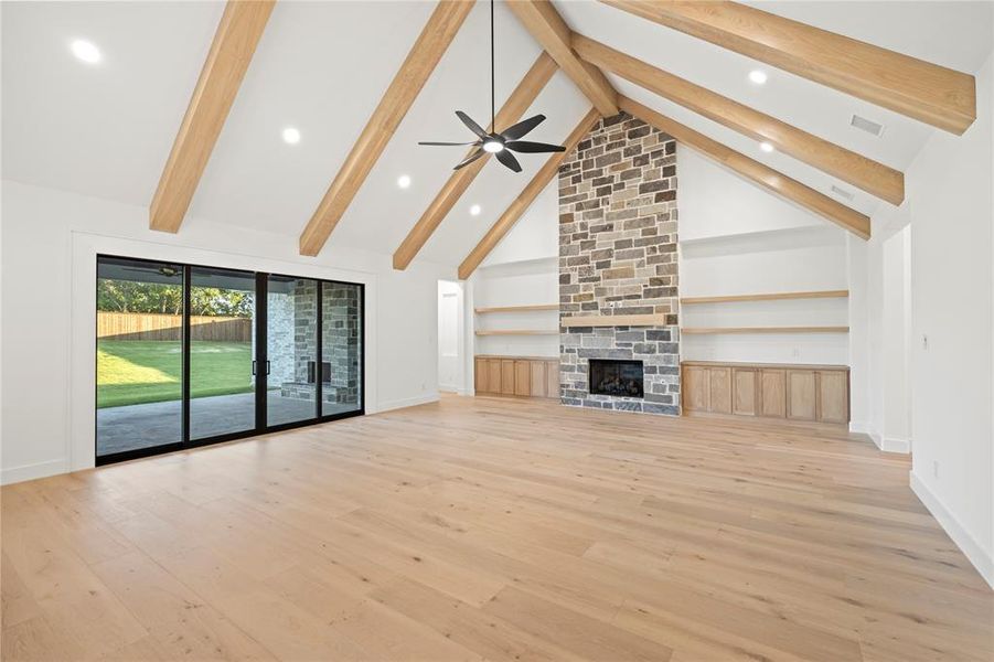 Vaulted ceiling with exposed wood beams, stone fireplace with a built-in hearth, wood-finish flooring, built-in shelving, and sliding glass doors opening to a concrete patio and manicured lawn