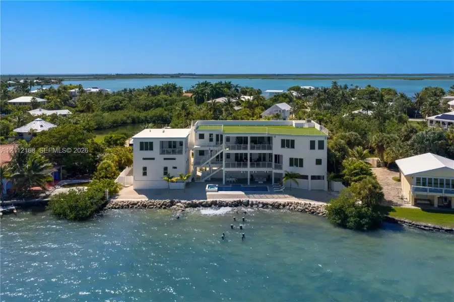 Waterfront multi-story residence featuring a ground-level swimming pool, expansive elevated balconies, stone-reinforced shoreline, and a rooftop deck Waterfront multi-story residence featuring a ground-level swimming pool, expansive elevated balconies, stone-reinforced shoreline, and a rooftop deck