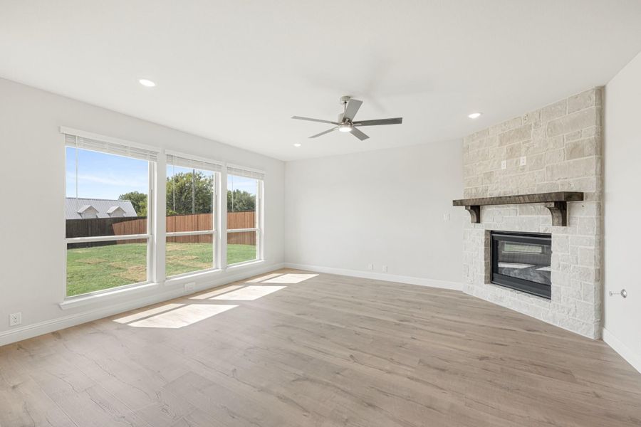 Open living room with hardwood floors, stone fireplace, ceiling fan, and large windows overlooking backyard