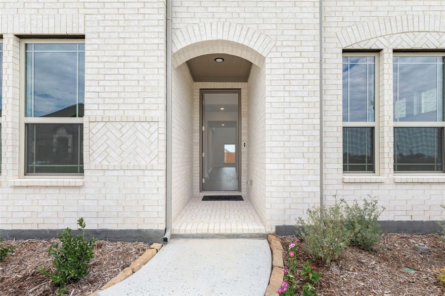 Front entry of a home with white brick exterior, arched doorway, gray door, and concrete walkway with landscaping.