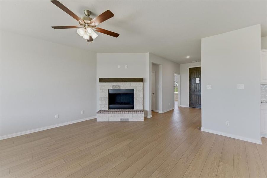 Expansive living area featuring light wood-finish flooring, a white brick fireplace with a wood mantel, a ceiling fan with integrated lighting, and a dark wood entry door with a glass insert Expansive living area featuring light wood-finish flooring, a white brick fireplace with a wood mantel, a ceiling fan with integrated lighting, and a dark wood entry door with a glass insert