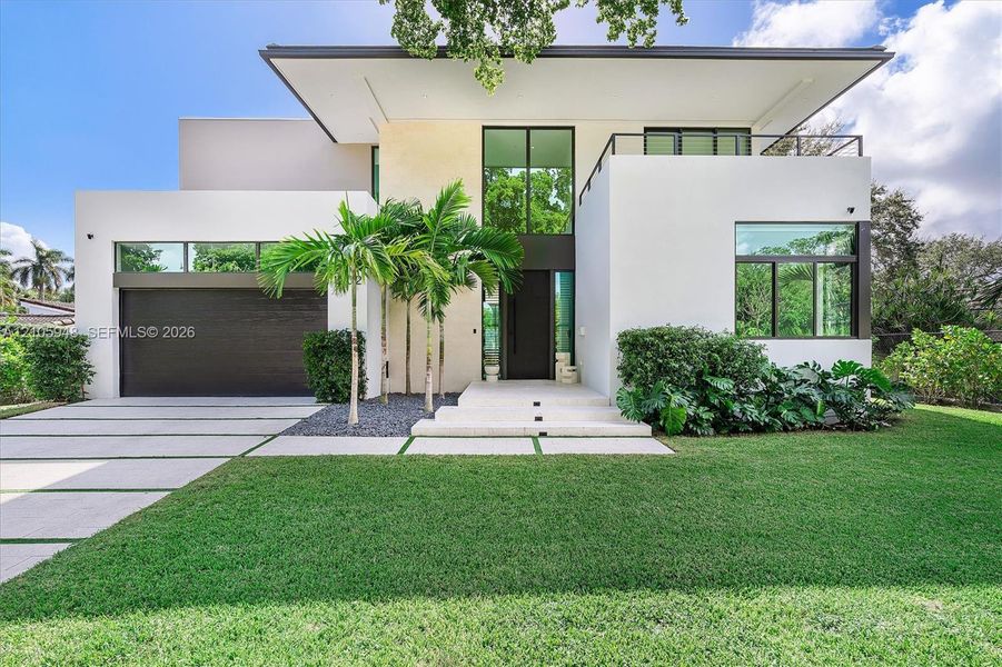 Modern architectural exterior featuring smooth stucco finish, geometric flat roofline, oversized floor-to-ceiling windows, second-story balcony with metal railing, and paver driveway with integrated turf strips