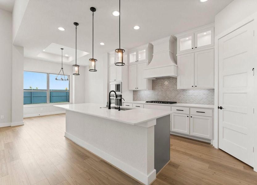 This modern kitchen features a spacious island, boasts ample cabinetry, a built-in oven, and a contemporary range hood. Pendant lights and recessed lighting illuminate the space, while large windows provide natural light and a view. The adjoining dining area enhances the open layout.