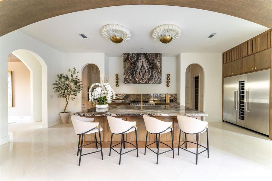 Kitchen with built in refrigerator, arched walkways, dark stone countertops, a kitchen breakfast bar, and brown cabinetry