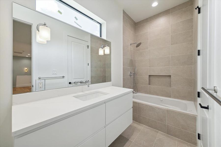 The ensuite bath for Secondary Bedroom No. 3 features an access door to the game room and a private door to the bedroom itself. It includes a tub/shower combo with designer grey tile walls and chrome plumbing fixtures, as well as an American Standard. Quartz countertops, designer porcelain floor tile, and a custom vanity with chrome cabinet hardware highlight the space. Additional features include a wall-mounted designer chrome faucet, an under-mount sink, a counter-to-ceiling mirror, and sconce lighting.