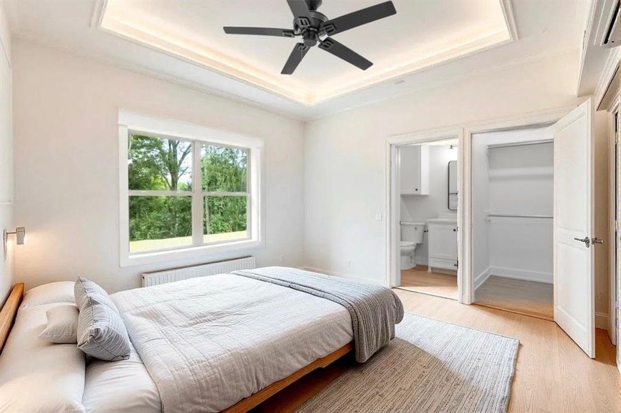 Bedroom with a tray ceiling, radiator, light wood finished floors, ensuite bath, and a ceiling fan