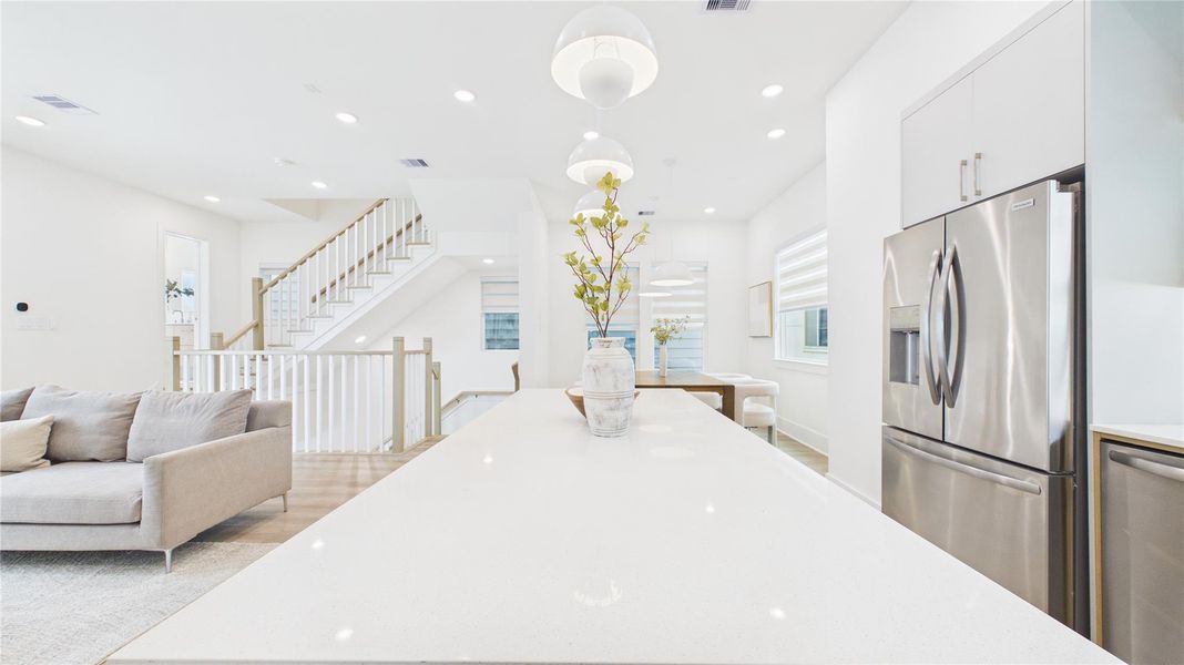 Bright, modern open-concept space featuring a sleek kitchen with stainless steel appliances, a large island, and elegant pendant lighting. Adjacent is a cozy living area with a staircase leading to the upper floor, creating a welcoming and spacious atmosphere.