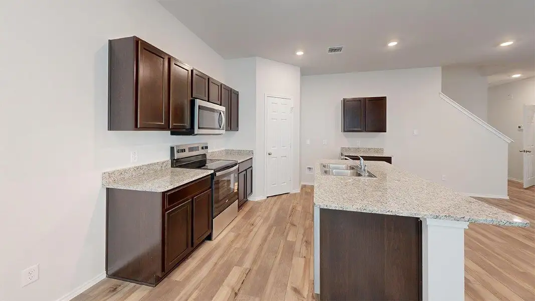 Kitchen featuring dark brown cabinetry, appliances with stainless steel finishes, a kitchen island with sink, light wood-style floors, and light stone counters Kitchen featuring dark brown cabinetry, appliances with stainless steel finishes, a kitchen island with sink, light wood-style floors, and light stone counters