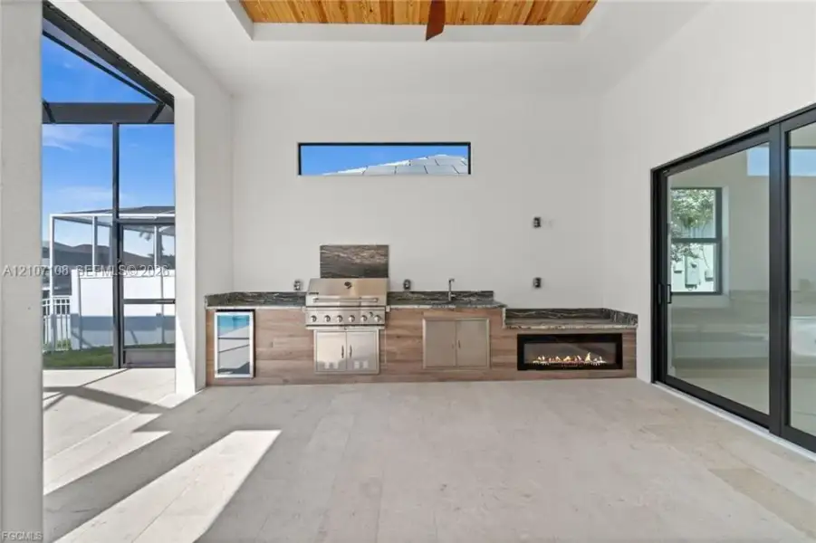 Covered lanai featuring a built-in outdoor kitchen with stainless steel appliances, stone-look countertops, wood-finish cabinetry, a linear fireplace, and tray ceiling with wood paneling