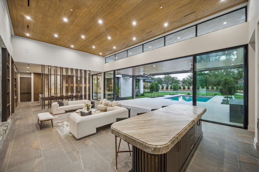 This elegant dining room is a masterclass in modern luxury, featuring Piasentina flamed natural tile flooring, designer pendant lighting, and in-ceiling speakers for a refined ambiance. Backlit, custom wallpapered built-ins add a dramatic touch, while floor-to-ceiling glass doors provide a stunning view and seamless indoor-outdoor flow.