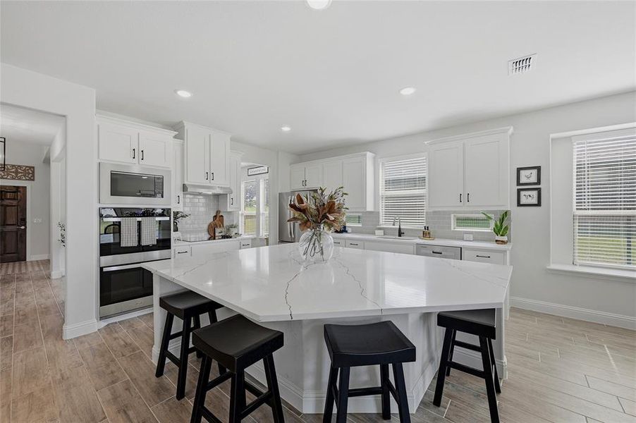 Spacious kitchen featuring an oversized island with countertop seating, white shaker cabinetry, stainless steel built-in appliances, a subway tile backsplash, and wood-finish flooring