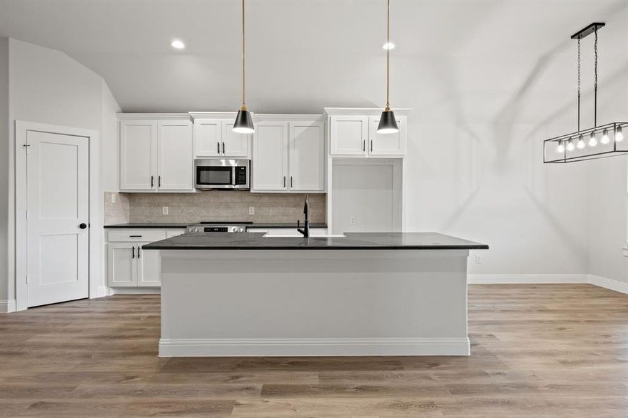 Kitchen featuring white cabinetry, hanging light fixtures, backsplash, an island with sink, and stainless steel microwave