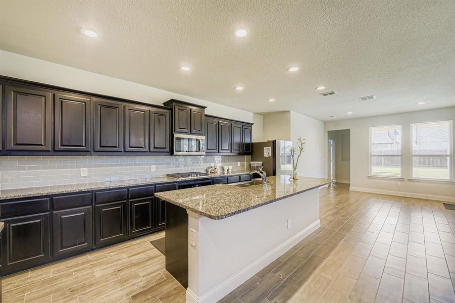 Kitchen with a textured ceiling, light stone counters, wood finish floors, tasteful backsplash, and a center island with sink Kitchen with a textured ceiling, light stone counters, wood finish floors, tasteful backsplash, and a center island with sink