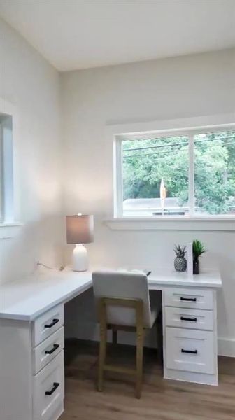 Room featuring built-in L-shaped desk with white cabinetry and dark hardware, two windows with white trim, and wood-finish flooring