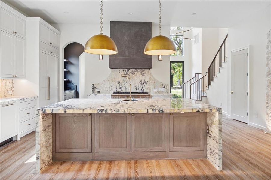 Close up view of the kitchen island with detailed waterfall marble countertops, custom brass fixtures and cabinetry for additional storage under the island, highlighted by overhead pendant lights. Double paneled Thermador dishwashers and paneled Thermador refrigertor and freezer columns to the left.