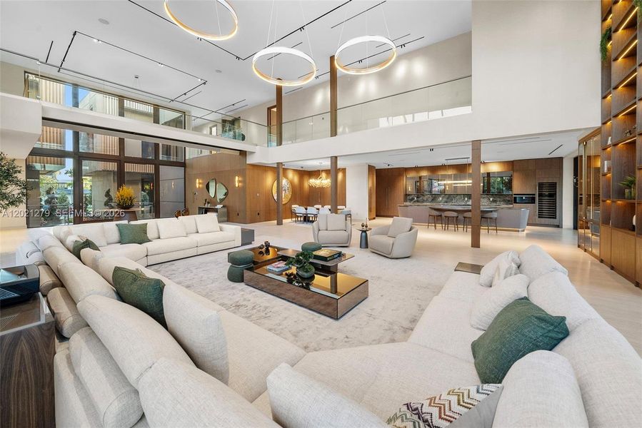 Double-height great room with triple ring chandelier, wraparound sectional seating, light stone floors, open second-floor glass balcony, walnut columns, full-height illuminated shelving, wine display, and open-plan dining and kitchen beyond.