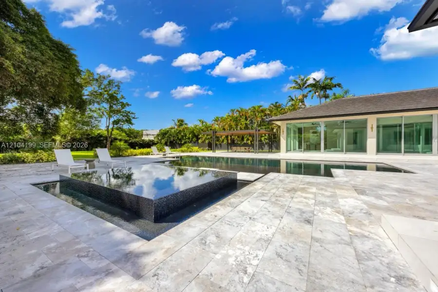 Backyard featuring a custom infinity-edge pool, a raised square spa with tile surfacing, extensive stone paver decking, and a covered pavilion structure