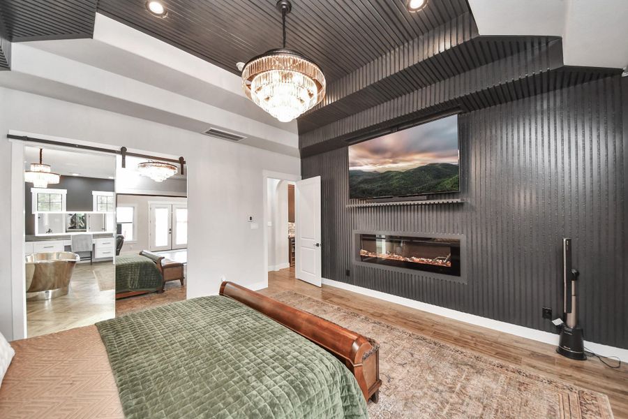 Experience luxury in this elegant bedroom featuring a sleek black accent wall with a modern fireplace and mounted TV. The room boasts a sophisticated chandelier, rich wood flooring, and a seamless transition to an opulent ensuite bathroom.