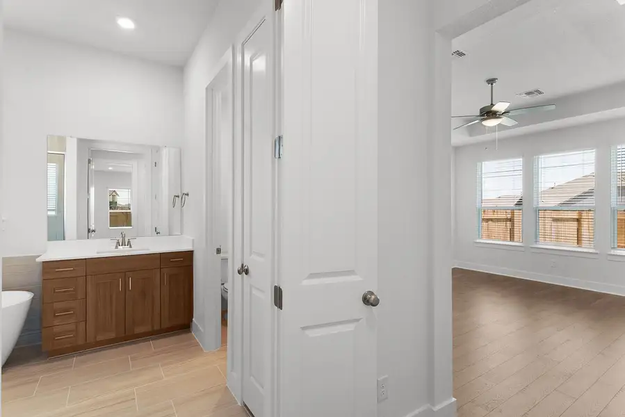 A well-appointed primary bathroom showcases a rich wood vanity and a refreshing shower, adjacent to a bright bedroom with sleek wood flooring.