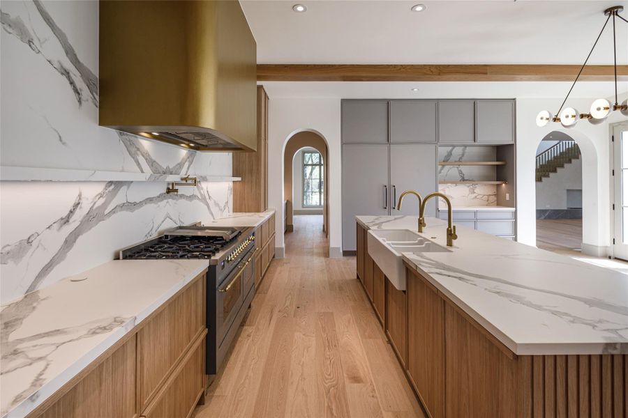 The materials do the talking. Dramatic veining wraps the counters and backsplash, a statement brass hood commands the range wall, and warm wood cabinetry keeps this brand-new kitchen from ever feeling cold.