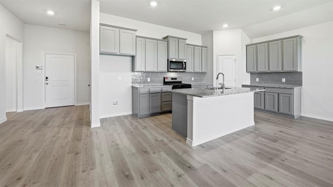 Kitchen featuring gray cabinetry, gas stove, an island with sink, light stone countertops, and stainless steel microwave Kitchen featuring gray cabinetry, gas stove, an island with sink, light stone countertops, and stainless steel microwave