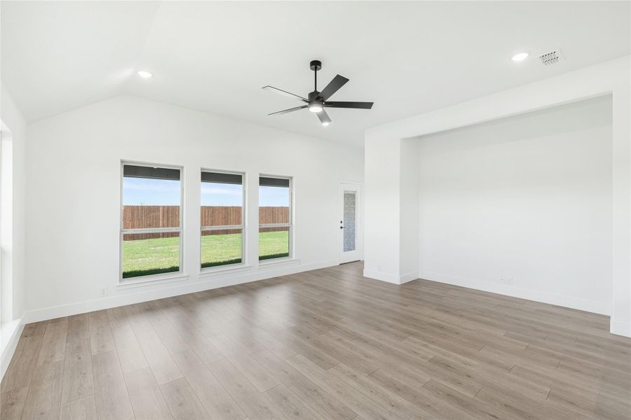 Open living room with vaulted ceiling, ceiling fan, light wood floors, and three backyard-facing windows