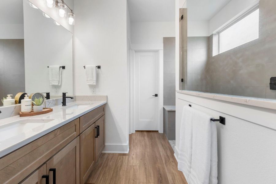 This bathroom features a modern design with a spacious double vanity, sleek black fixtures, and a large mirror. The room is bright with natural light from a high window, and it has a mix of wood and tile flooring for a contemporary look.