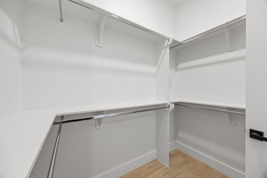This photo depicts a spacious walk-in closet featuring multiple hanging rods and shelves, ideal for organized storage. The bright, neutral color scheme enhances the open feel, complemented by a light wood-like floor.