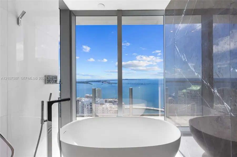 Freestanding soaking tub featuring chrome floor-mounted faucet, floor-to-ceiling windows with water backdrop, and large-format tile wall surrounds